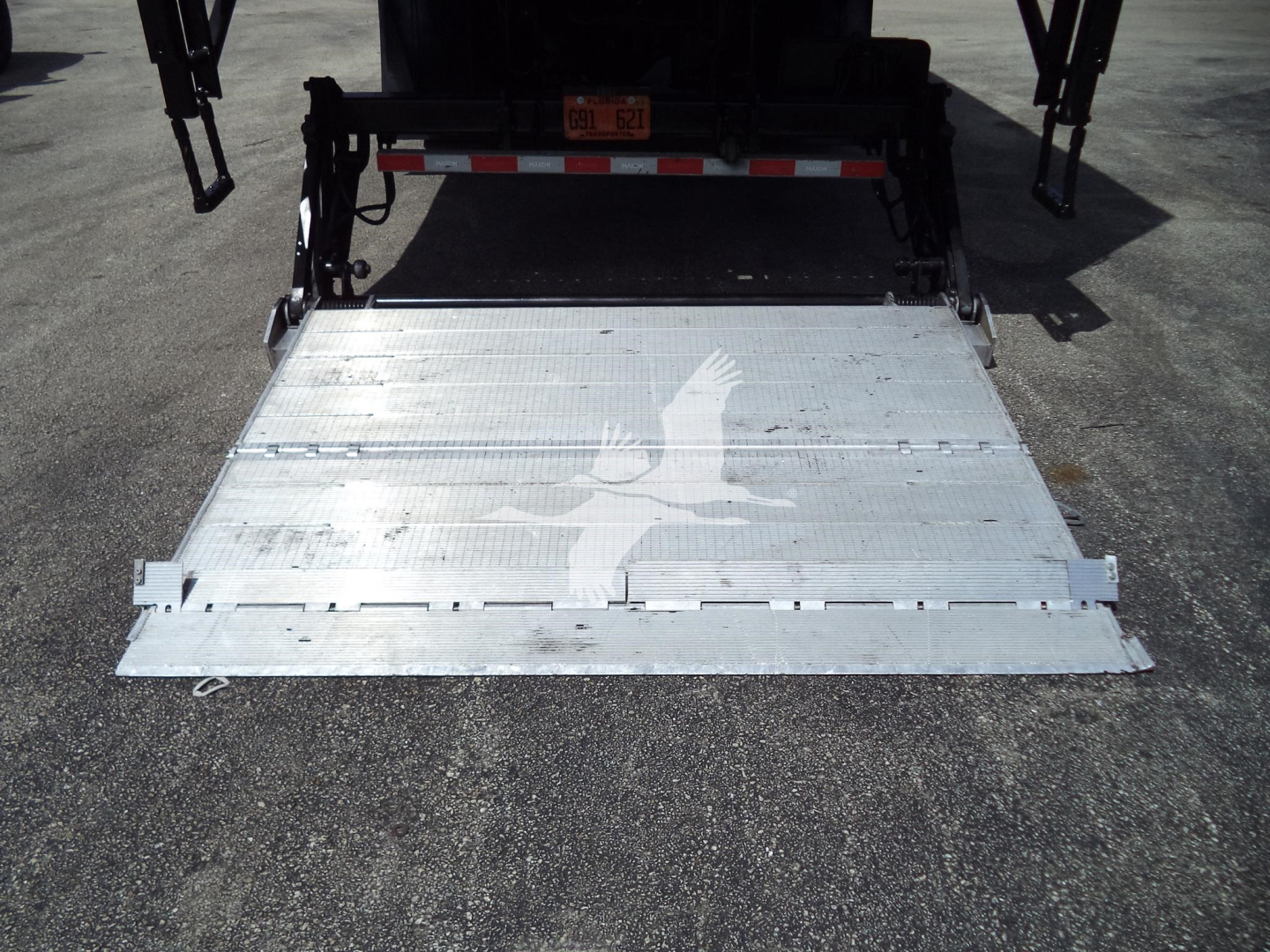 International MV power tailgate/roll-off bed showing aluminum loading ramp and deck with faint bird logo.