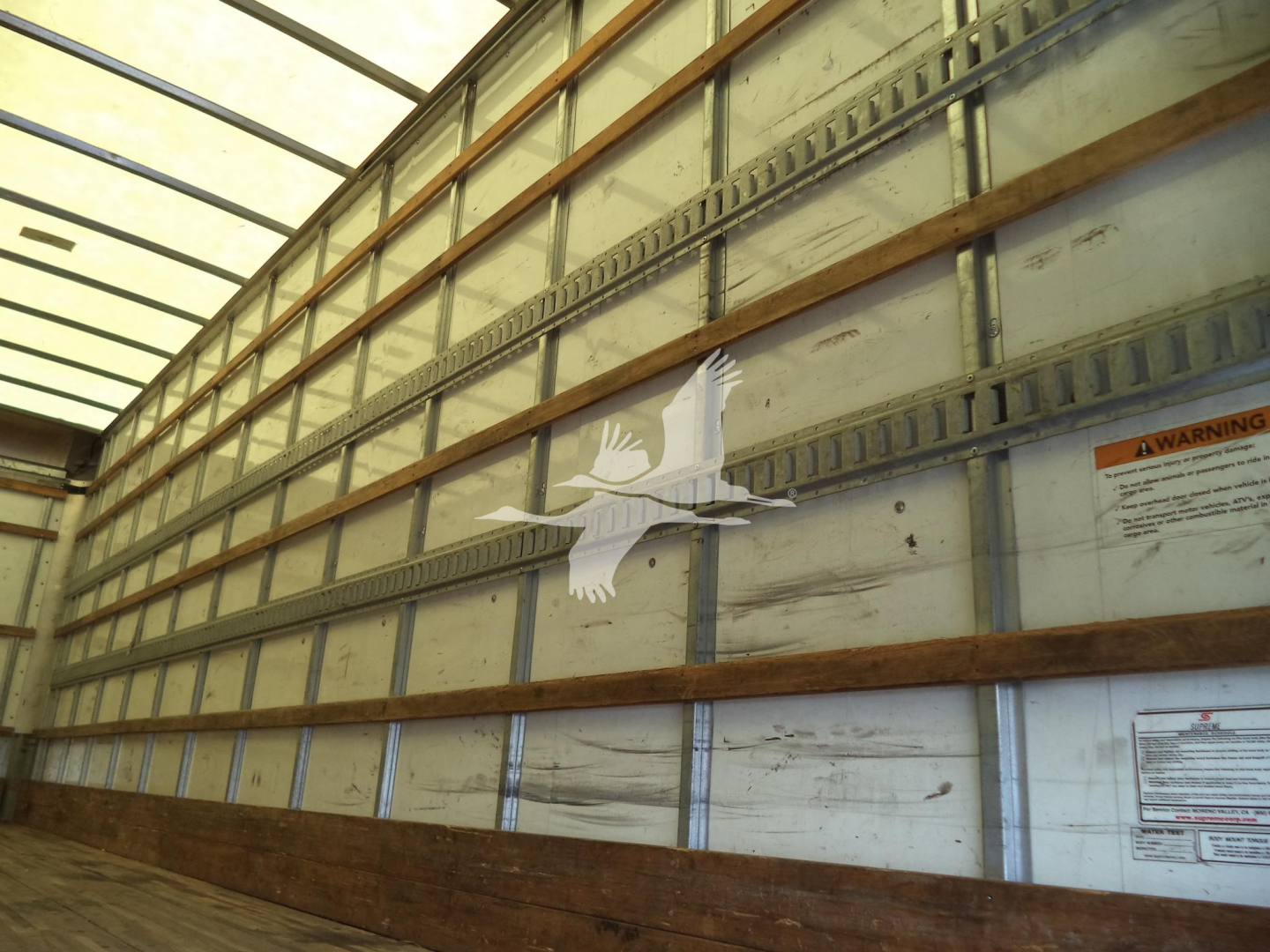 Truck trailer interior with metal walls and wooden runner slats, warning label on side, empty cargo space.
