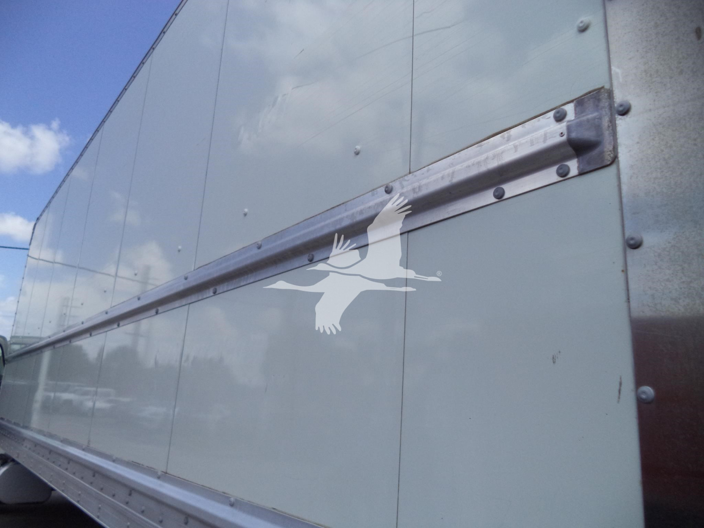 Truck trailer side showing riveted metal panels and horizontal aluminum rail; window reflection on light gray surface.