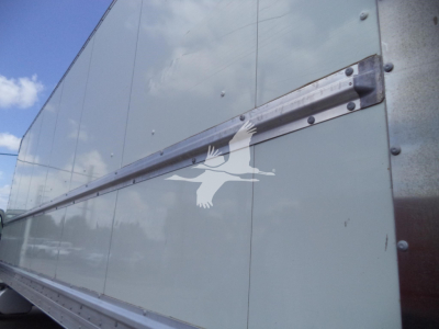 Truck trailer side showing riveted metal panels and horizontal aluminum rail; window reflection on light gray surface.