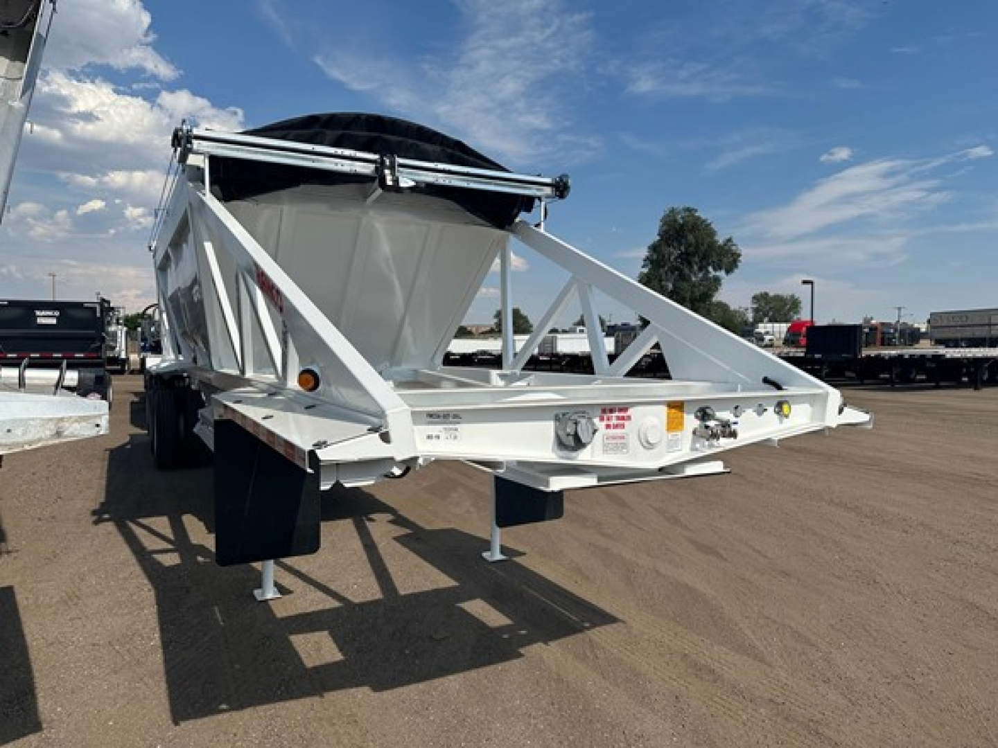Bottom dump trailer in a parked lot, white steel frame with a large steel hopper and a black cover on top; front hitch area visible with support legs deployed.