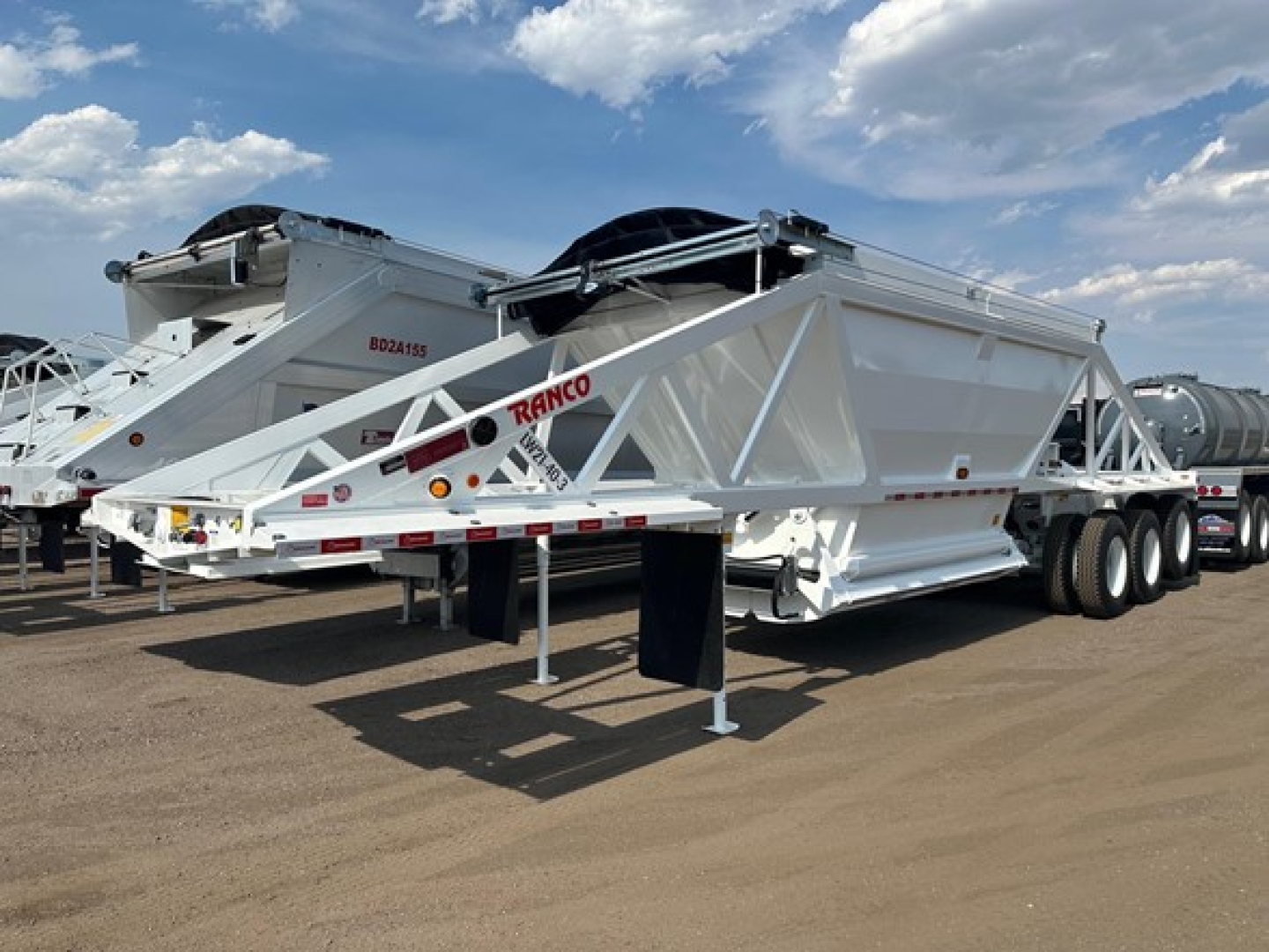 Ranco tri-axle bottom dump trailer in a parking lot, white with red logos. Visible text includes 'RANCO' on the frame and a small serial/ID marking, with a spare tire mounted on the upper area.