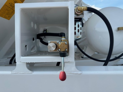 Dump trailer valve block inside a square white compartment with a brass gate valve and red-tipped lever, hydraulic hose and propane tank visible.