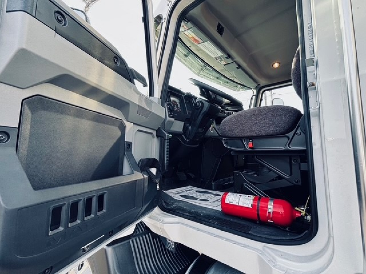 Cab interior of a 2025 Mack MD7 truck showing driver seat, steering wheel, and open door with a fire extinguisher on the floor.