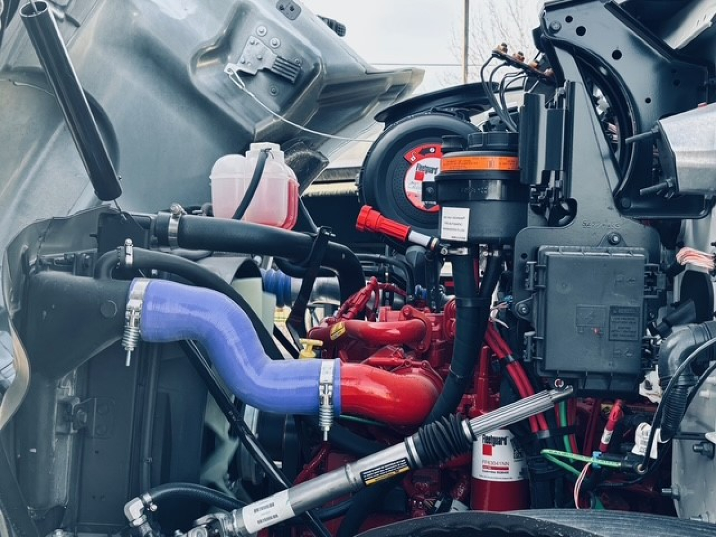 Mack MD7 engine bay view showing blue coolant hose, red intake manifold, black wiring harness, pink reservoir, and various engine components.