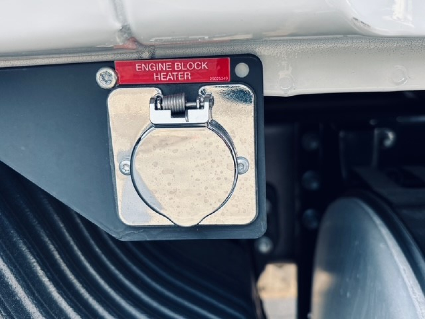 Engine block heater cover with red label reading 'ENGINE BLOCK HEATER' and circular electrical plug housing on a Mack MD7 engine bay.