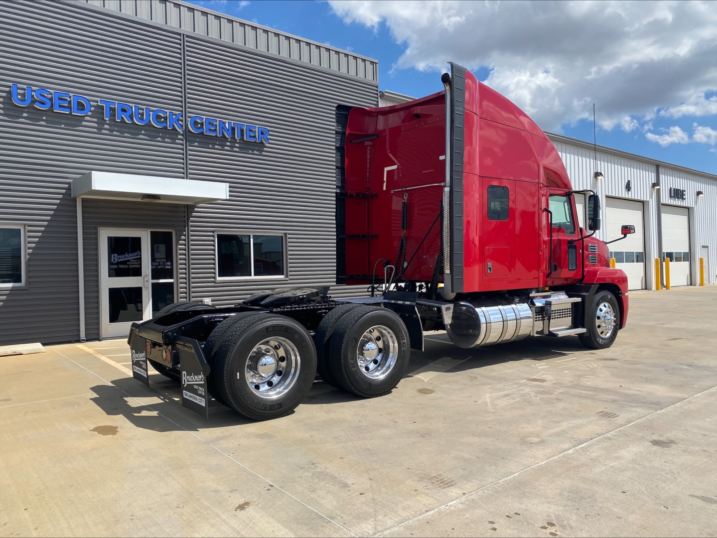 Truck in red cab with black chassis and dual rear axles parked outside a Used Truck Center building.