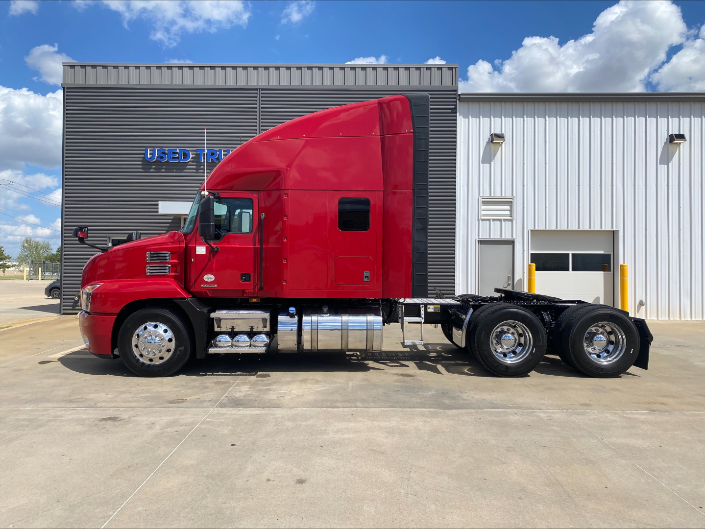 2020 Mack AN64T70 tractor in red side view showing sleeper cab and dual rear axles, chrome fuel tanks, and bannered dealership building in background.