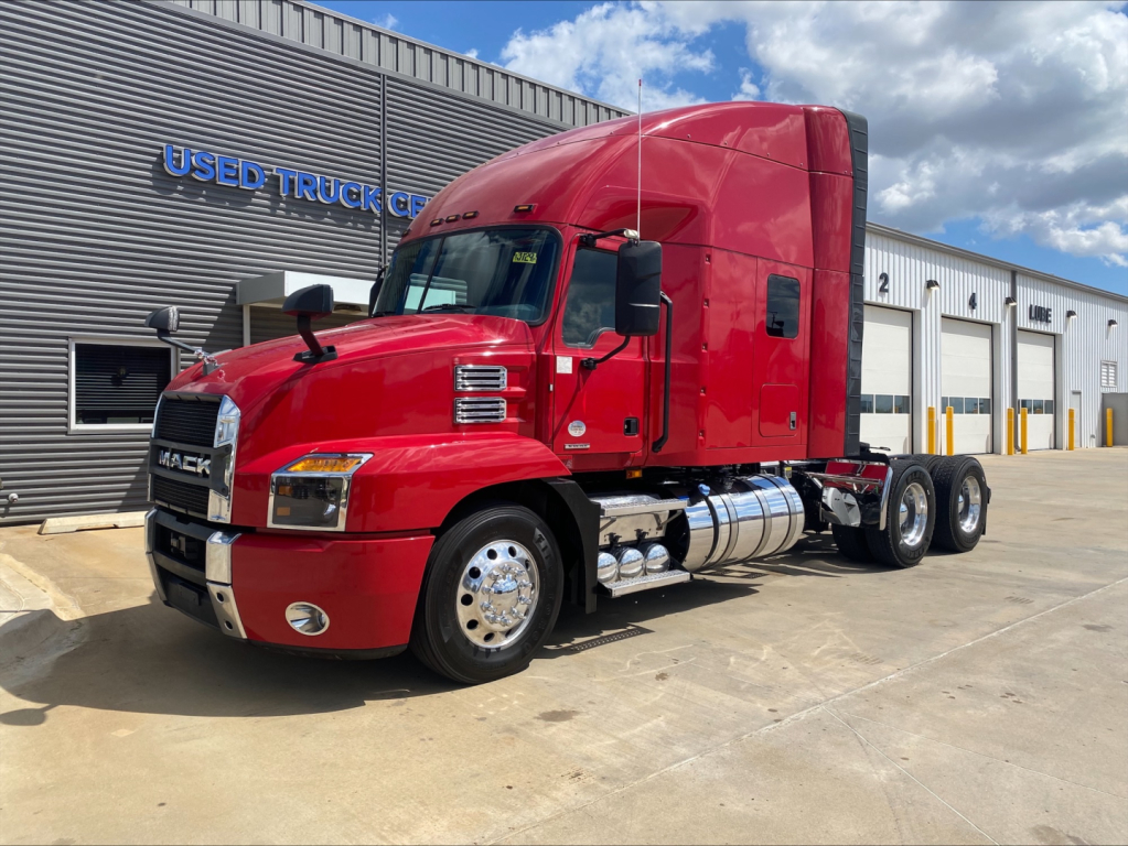 2020 Mack AN64T70 tractor with red cab and sleeper in front of a service bay, showing side view and chrome fuel tanks.