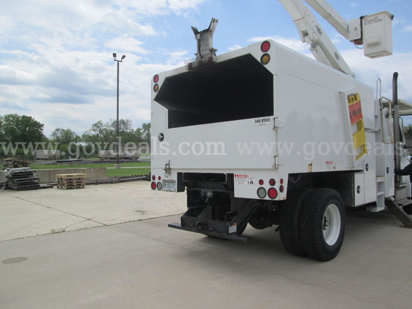 Back view of a white Freightliner M2 106 bucket truck with open service bed, rear bumper, warning labels, and boom crane mounted on right side.