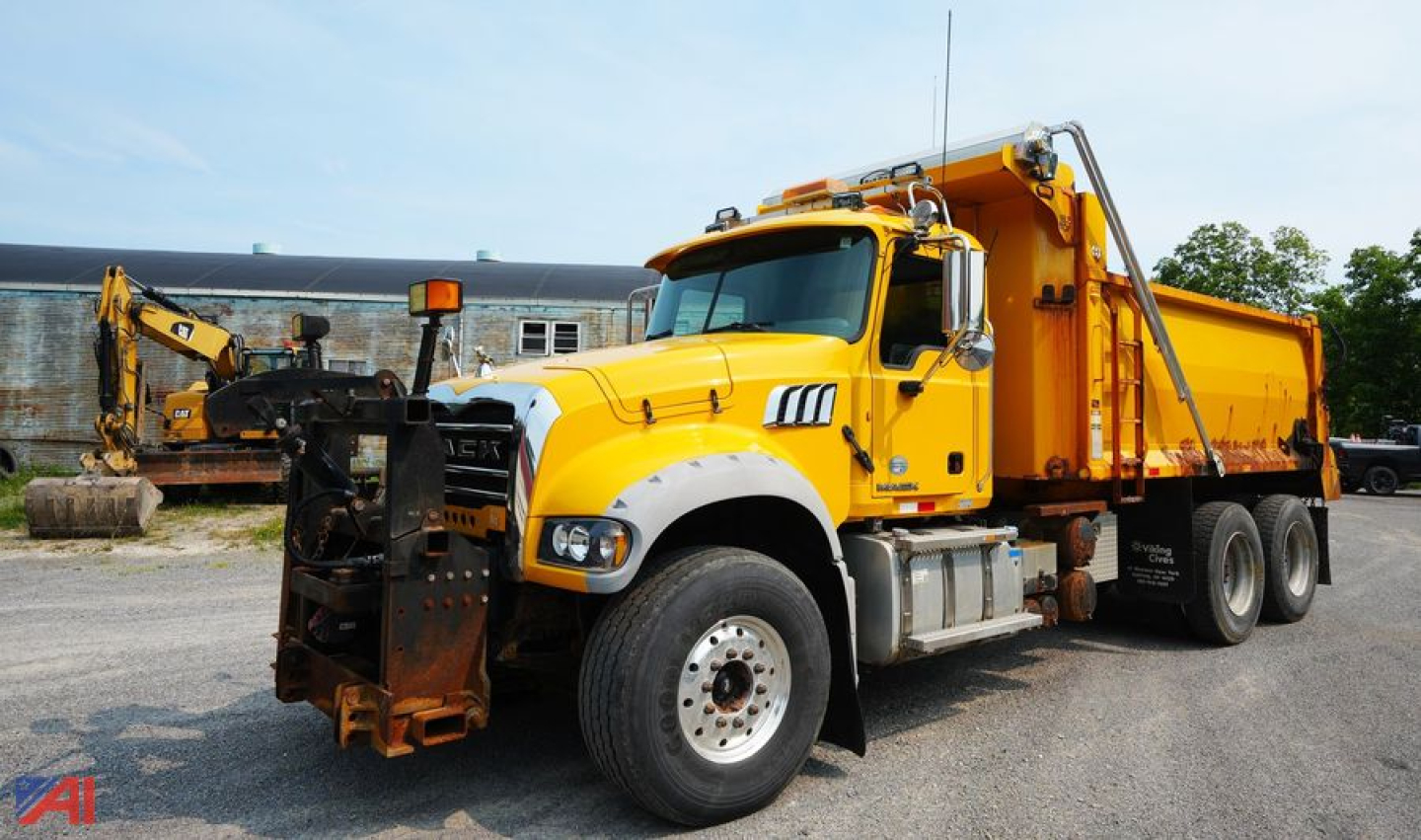 Dump truck front-left view of a yellow 2014 Mack GU713 dump truck with plow attachment visible, cargo bed in background, daytime outdoor lot.