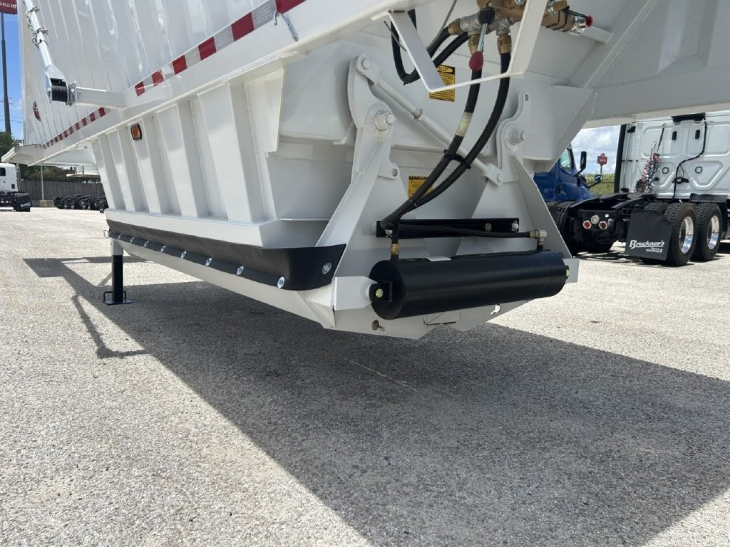 Dump trailer underside showing landing gear, hydraulic hose, and black undercarriage protector; white steel chassis and part of the dump bed with reflective stripes