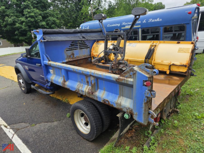 Dump truck bed with mounted plow equipment and RAM 4500 style cab visible; blue dump bed shows rust and wear; yellow salt spreader or hopper mounted behind cab; vehicle in a parking lot.