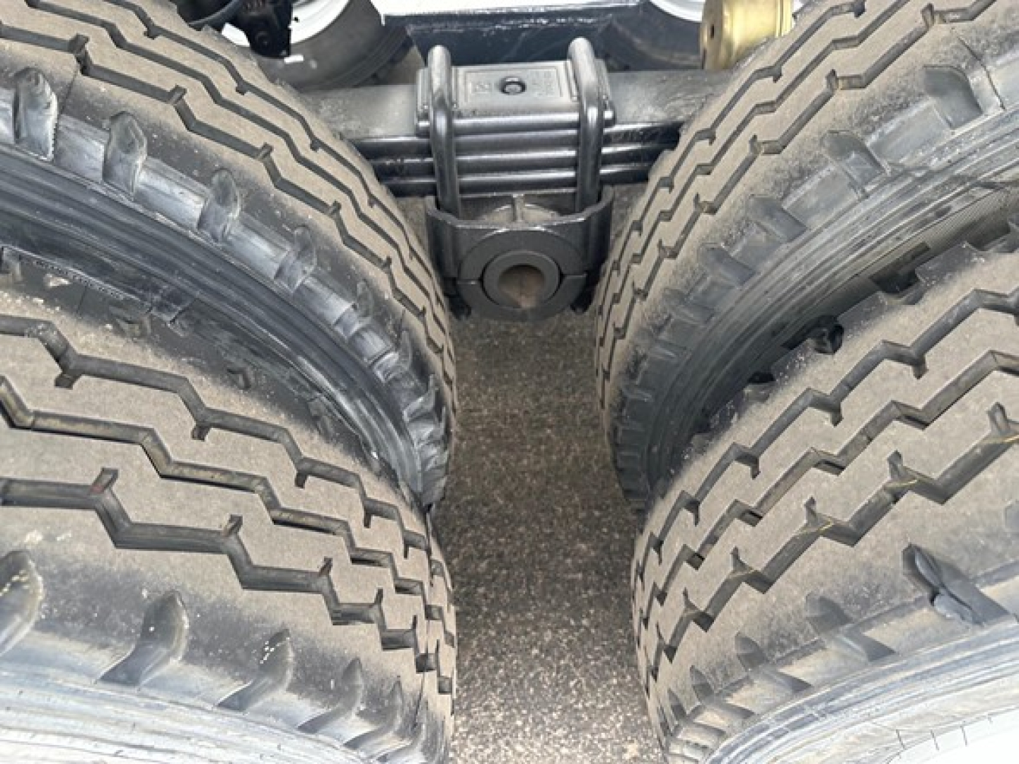 Trailer axle setup showing dual tandem wheels and suspension, with heavy-duty leaf springs and connecting pin; no visible text.