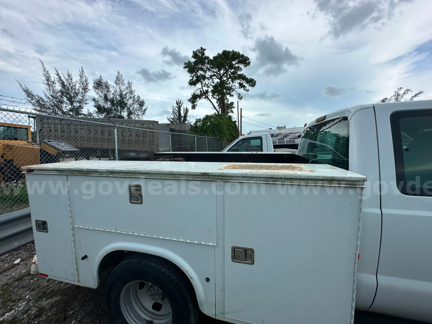 Ford F-350 service body truck side view with storage compartments and tool boxes, white utility bed, cab visible on right, open truck yard in background.