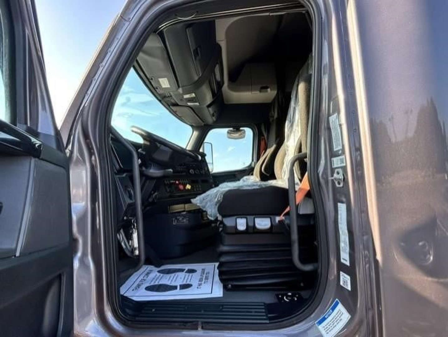 Truck cab interior view of a 2023 Freightliner Cascadia 126 sleeper showing steering wheel, dashboard, and driver's seat from open door.