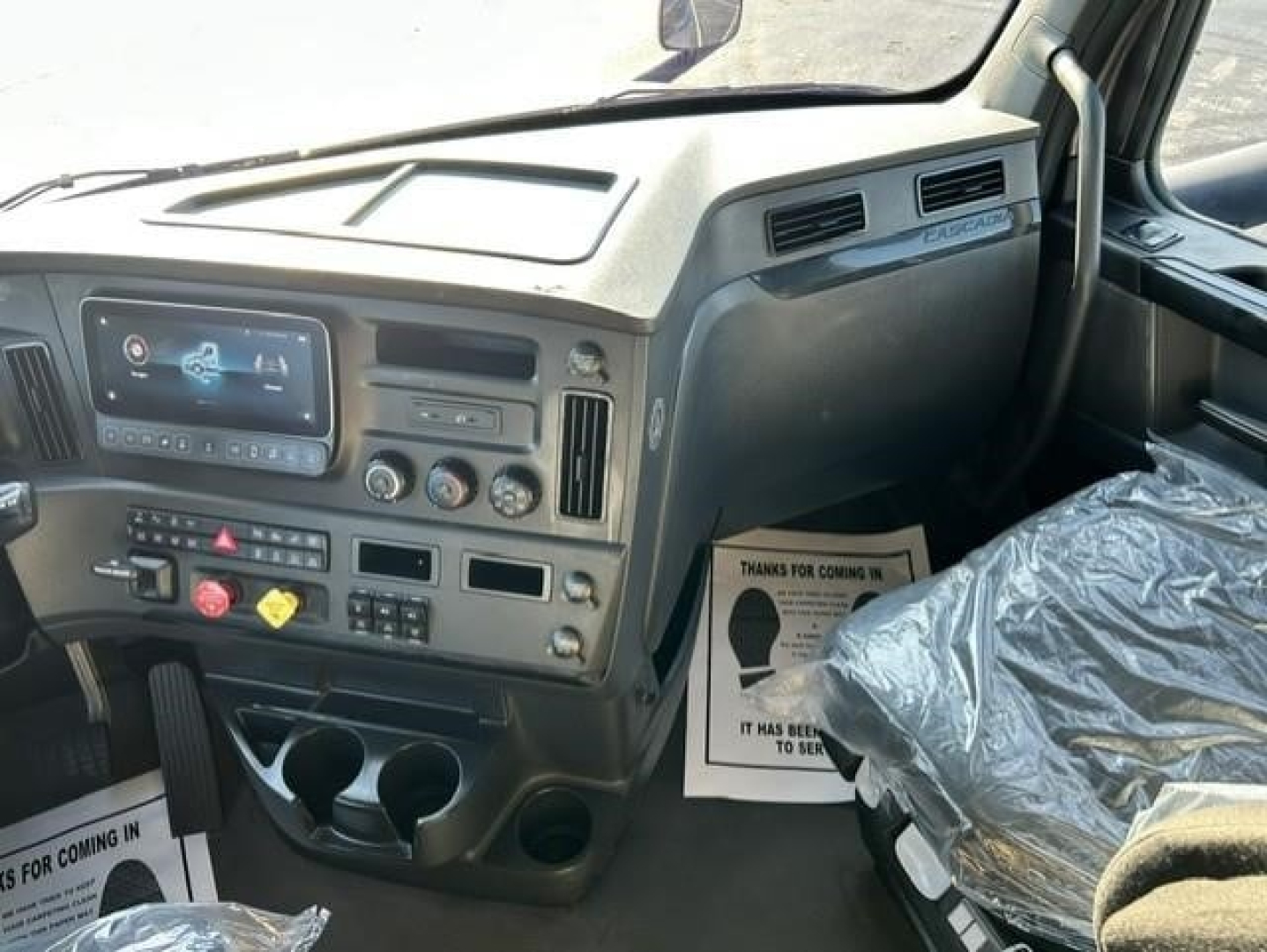 Dashboard of Freightliner Cascadia interior showing steering column, infotainment screen, climate controls, and storage cups; plastic seat covers and floor mats visible.