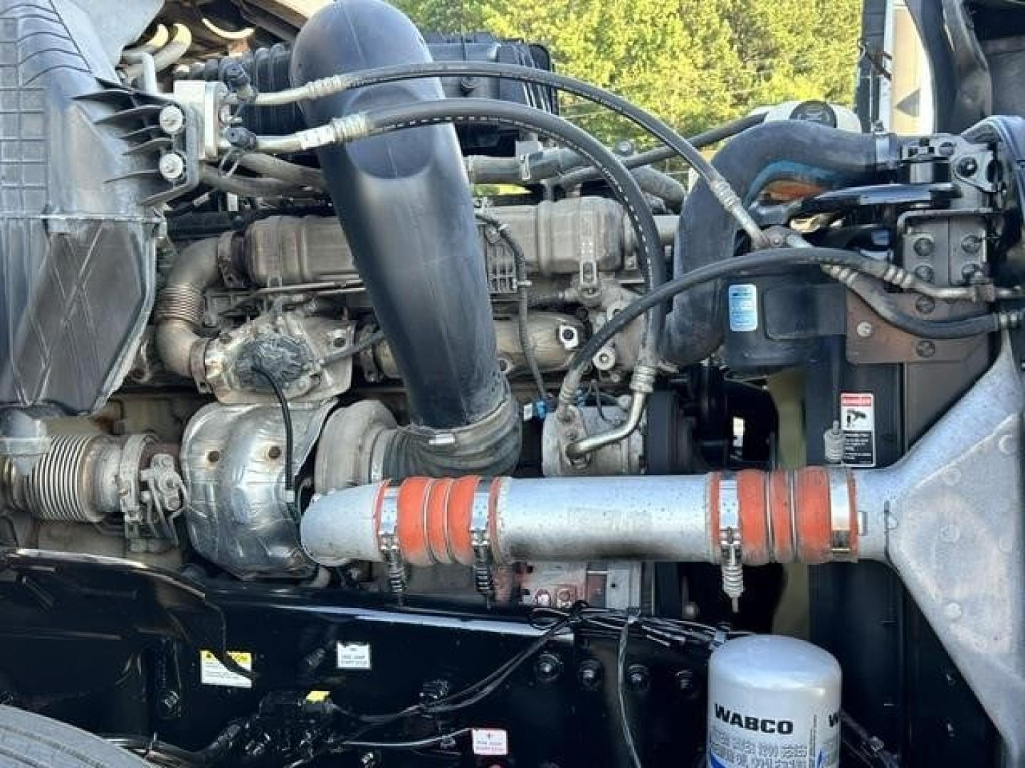 Engine bay of a Freightliner Cascadia 126 showing hoses, intercooler piping, turbo, radiator intake, and wiring with a silver pipe strapped by orange clamps.