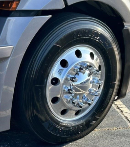Truck front wheel close-up showing chrome polished hub, tire, and rim on Freightliner Cascadia 126. Visible tire sidewall branding and wheel lug nuts.