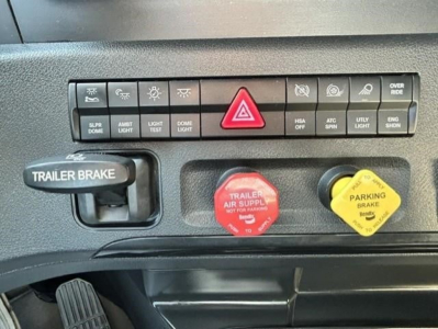 Dashboard panel of a Freightliner Cascadia showing hazard button, light controls, trailer brake handle, red air supply knob, and yellow parking brake knob.
