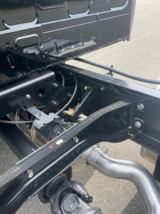 Front chassis area of a 2027 Hino L7 cab chassis truck showing frame rails, wiring, hoses, and exhaust/hood components with black metal surface.