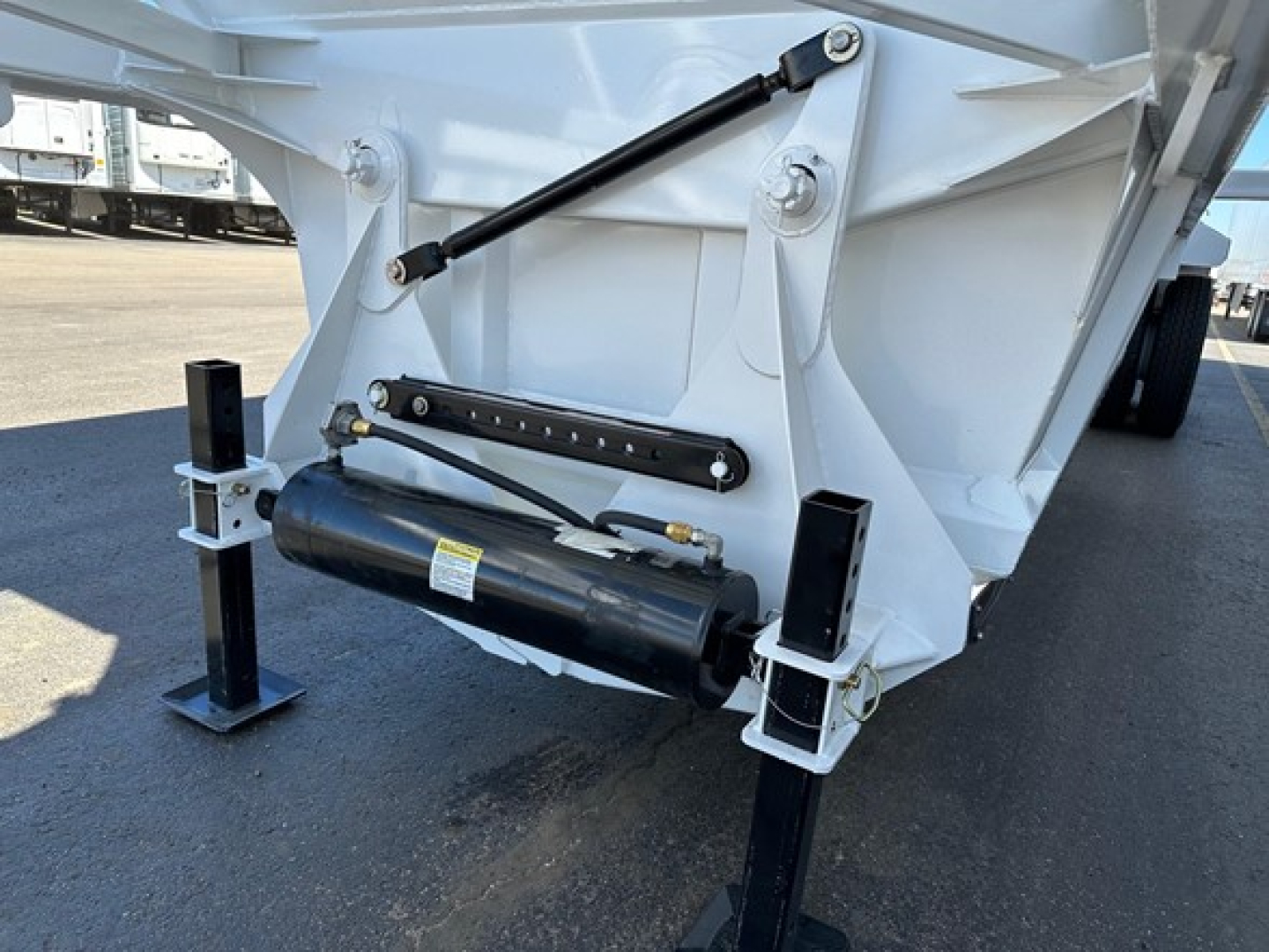 Bottom-dump trailer hitch area showing hydraulic cylinder and support legs with white painted frame and black components.
