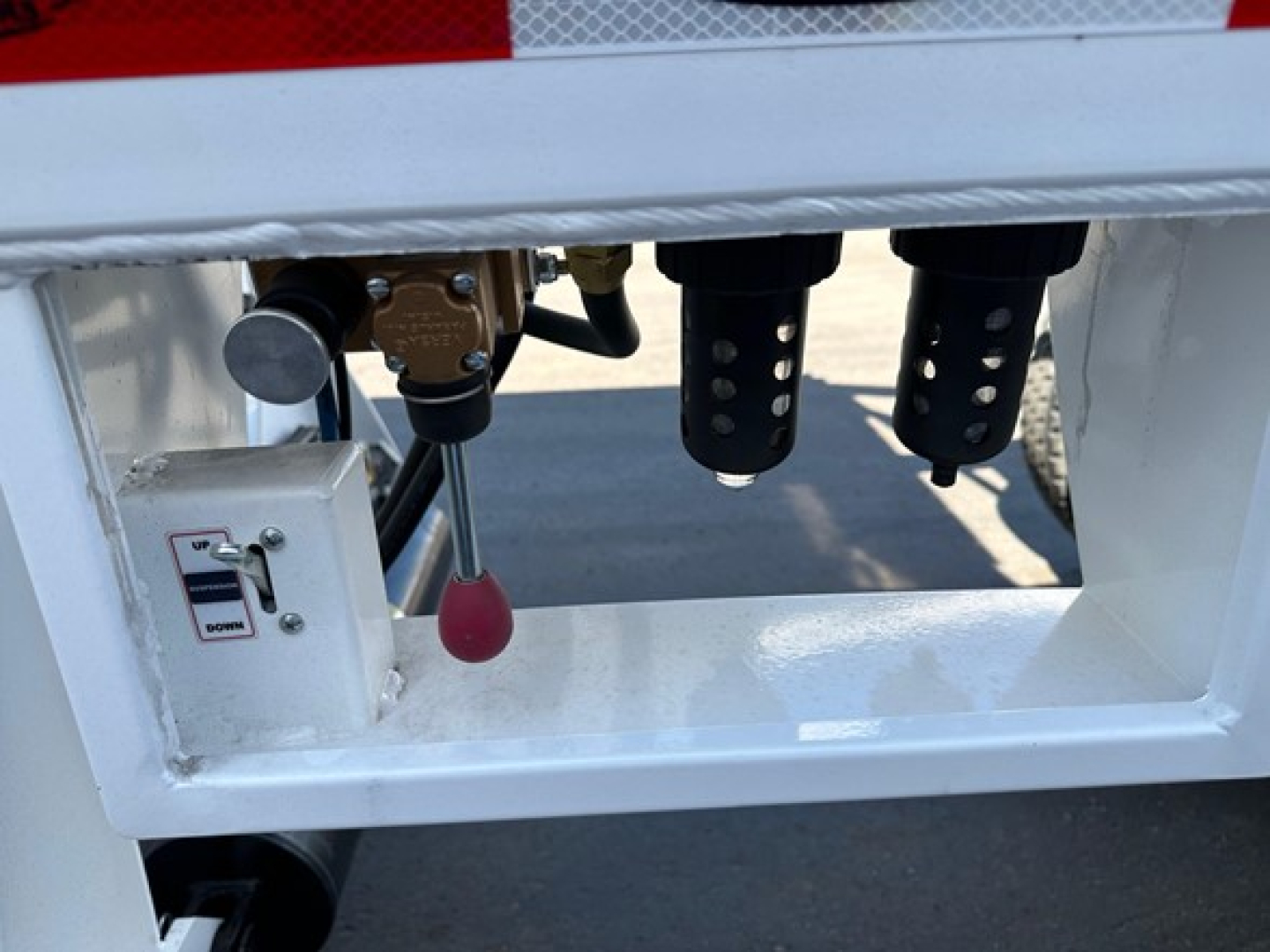 Dump trailer undercarriage view showing control lever and valve box with labels, hydraulic line fittings, and black suspension or mounting components.