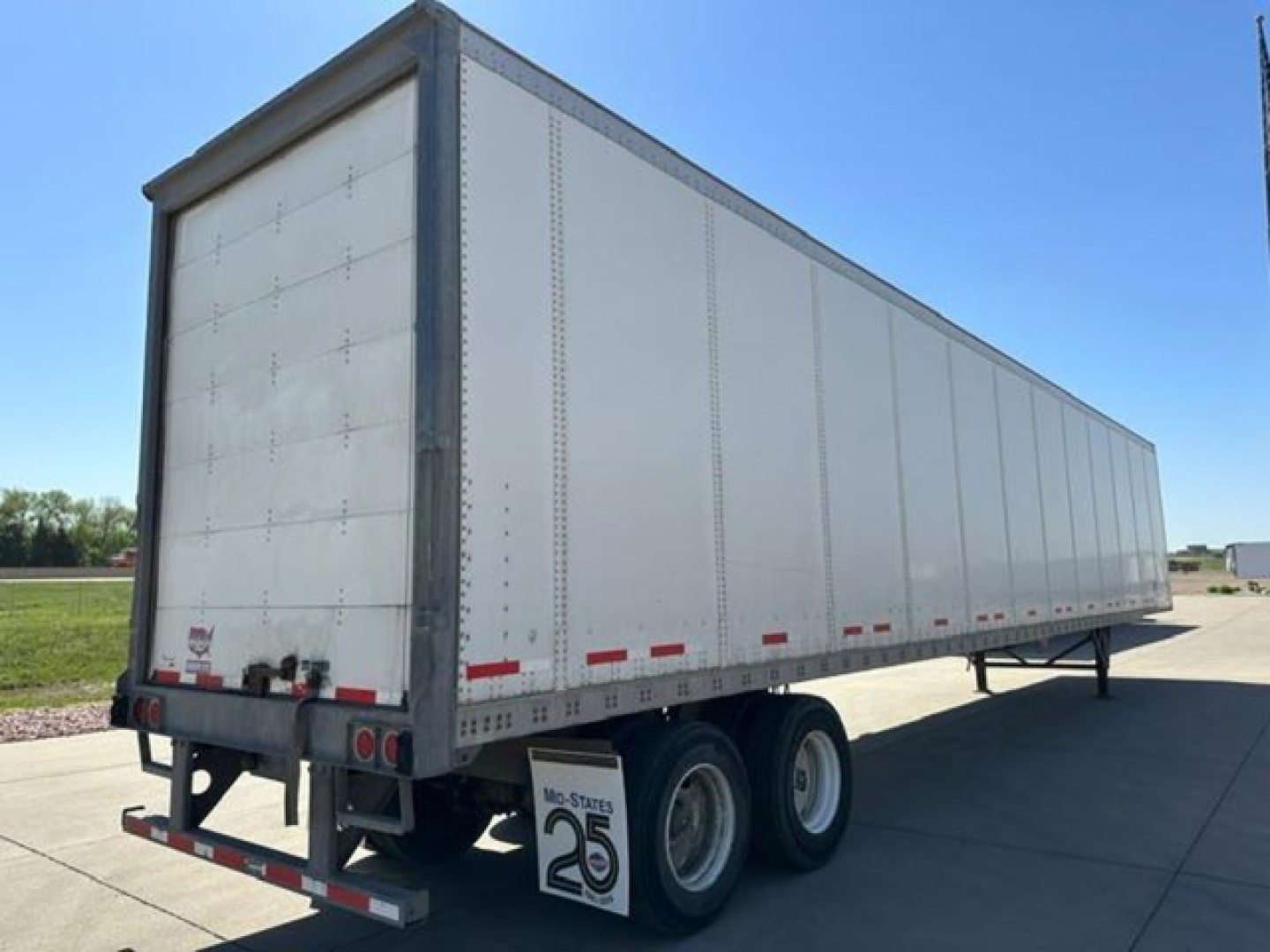 Trailer showing a long white box van with tandem rear axles and wheels, viewed from side at an open lot.