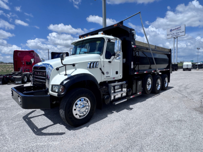 Dump truck with white cab and black dump body, Mack GR84F, front grille, dual side mirrors, three rear axles visible, loader. Visible text includes Mack emblem on grille and truck accessories.