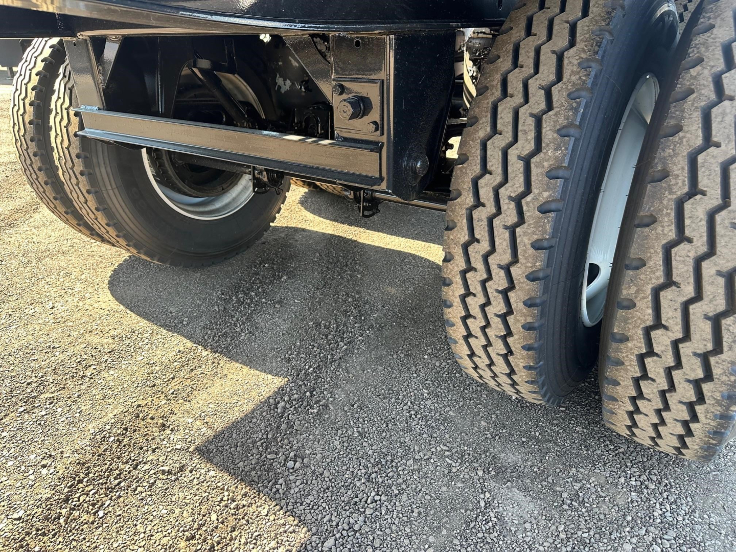 Trailer undercarriage showing dual wheels and suspension with metal frame and leaf spring; visible tires and gravel ground.