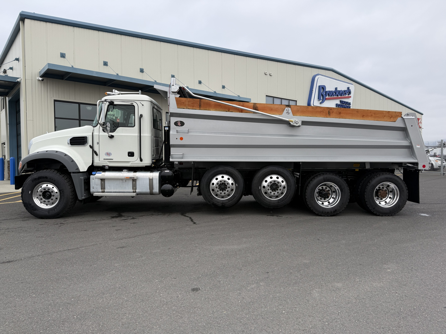Dump truck side view of a 2025 Mack GR104F with a white cab and silver dump bed parked in front of a building.