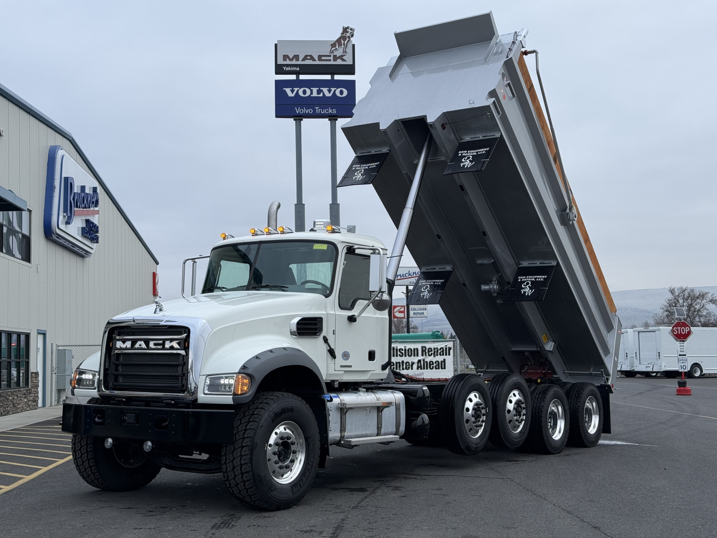 Mack GR104F dump truck with tilting bed raised, white cab, black grille, multiple rear axles, stickered side mirrors, dealership lot in background.