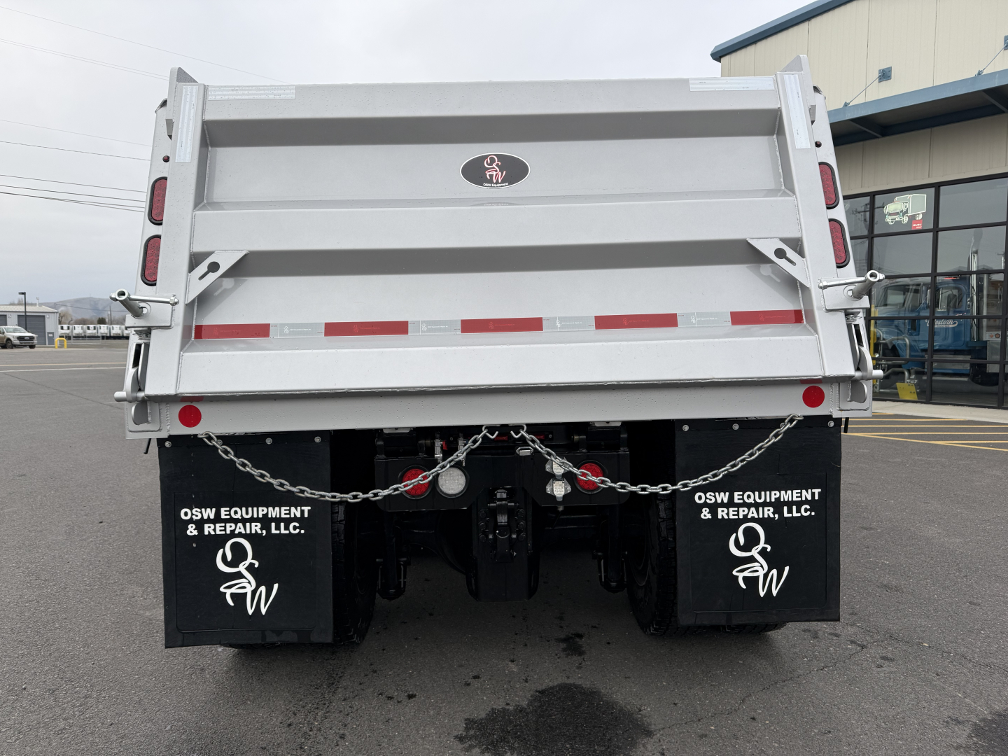 Dump truck bed rear showing OSW Equipment & Repair, LLC mud flaps, taillights, chains, and rear hydraulic connections on a Mack GR104F with a silver dump body.