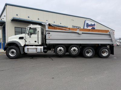 Dump truck side view of a 2025 Mack GR104F with a white cab and silver dump bed parked in front of a building.