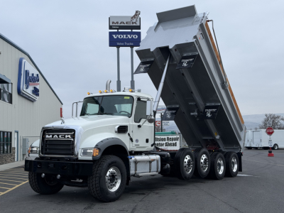 Mack GR104F dump truck with tilting bed raised, white cab, black grille, multiple rear axles, stickered side mirrors, dealership lot in background.