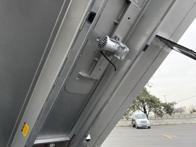 Dump truck component close-up showing a motorized device attached to a metal panel, with labels and wiring visible.