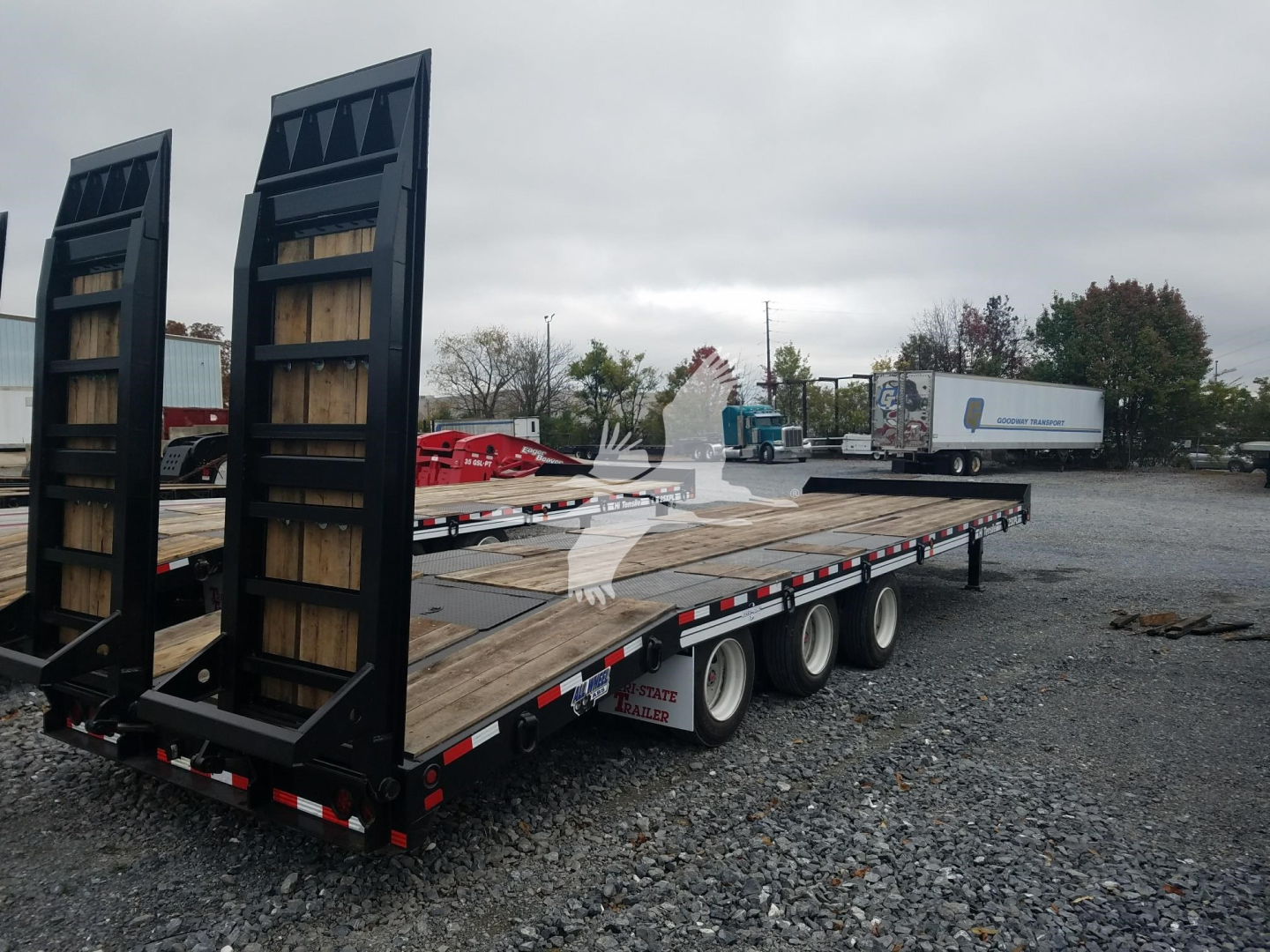 Flatbed trailer showing rear ramps and wooden deck with dual rear axles and gravel lot.