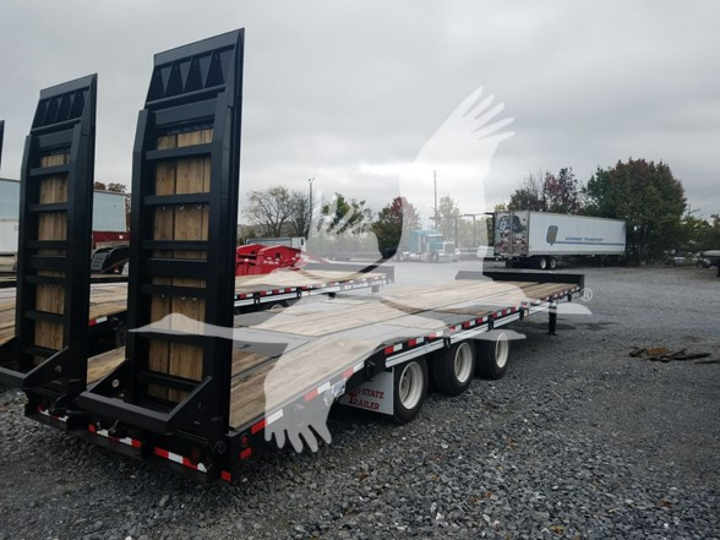 Trailer flatbed with ramp ends and multiple axles in a gravel lot, showing wooden deck and metal frame.