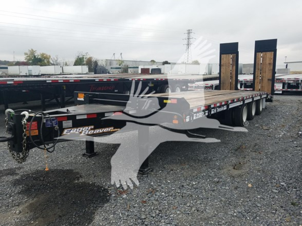 Flatbed trailer with dual goosenecks and wood deck, Eager Beaver logo visible, chain and hitch at front, gravel lot with other trailers in background.