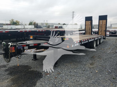 Flatbed trailer with dual goosenecks and wood deck, Eager Beaver logo visible, chain and hitch at front, gravel lot with other trailers in background.