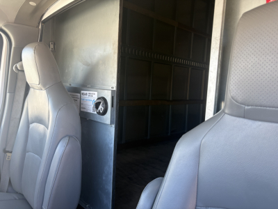 Ford E-350 cargo van interior showing partition wall and seating in foreground; metal cargo area with panels and taped label on wall.