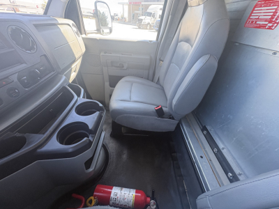 Ford E-350 cargo van interior showing front seats and dashboard with cup holders; steering wheel and driver side door; red fire extinguisher lying on the floor near the center; VIN and stock details not shown in image.