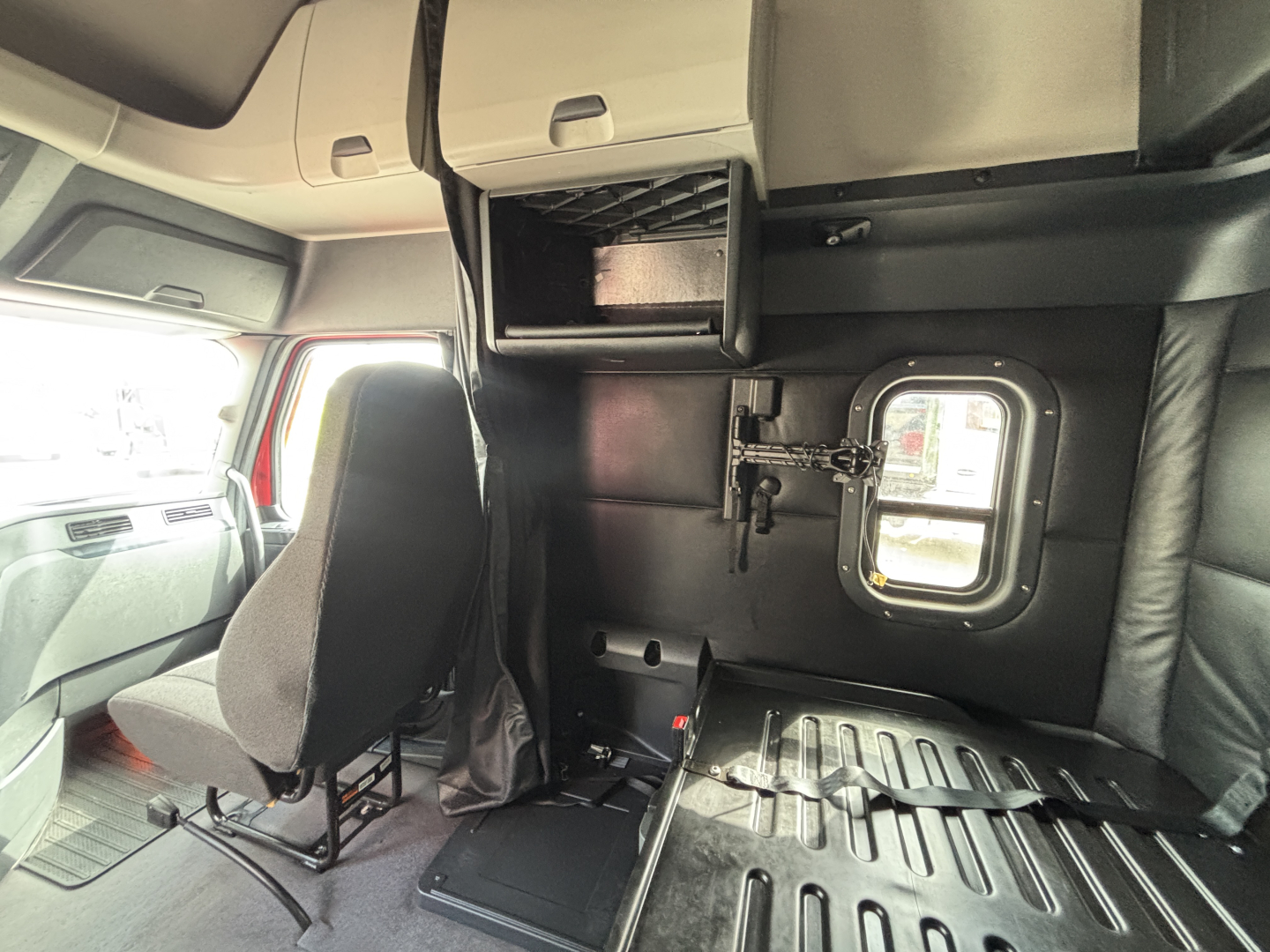 Truck cab interior showing storage compartments and sleeper area with window hatch and seat; cabin walls with a small closet-like box above and a metal floor plate.