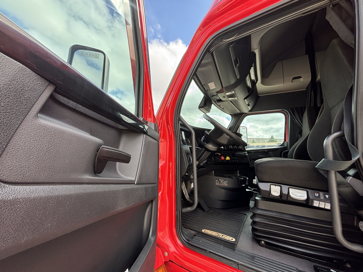 Truck cabin interior view from open driver door showing steering wheel, dashboard, pedals, and seat with red exterior.