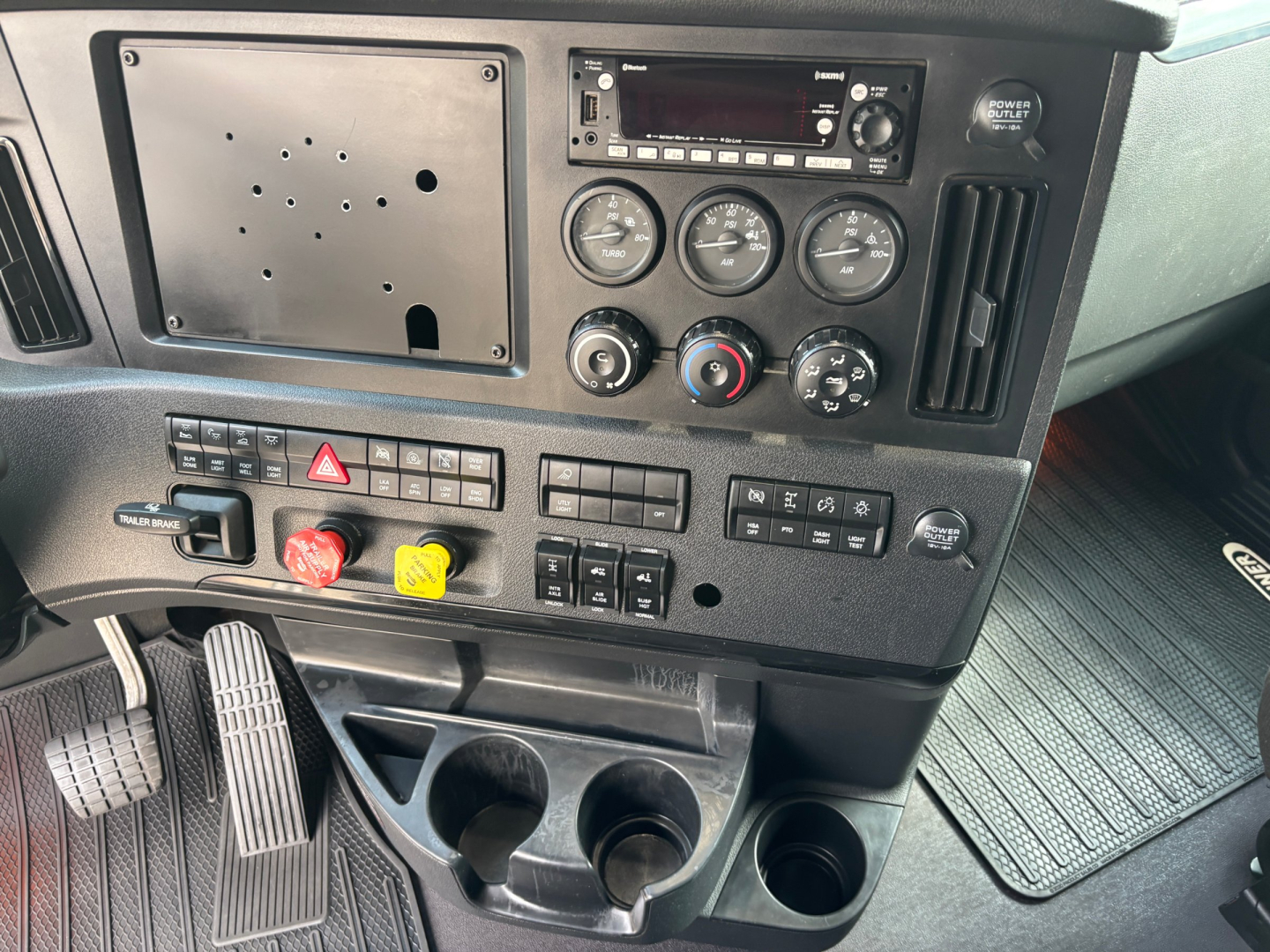 Dashboard and control panel of a Freightliner truck showing gauges, radio, and control switches; three round gauges, hazard lever, climate controls, and cup holders.