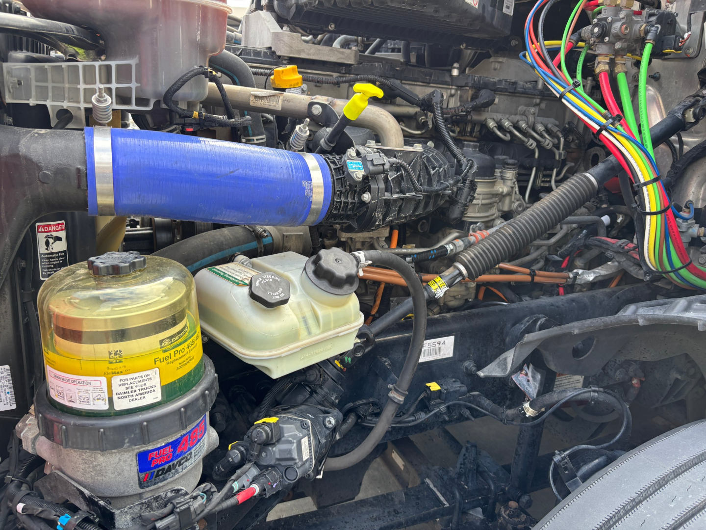 Engine bay showing blue intercooler hose, coolant reservoir, fuel filter, and wiring harness in Freightliner truck engine bay.