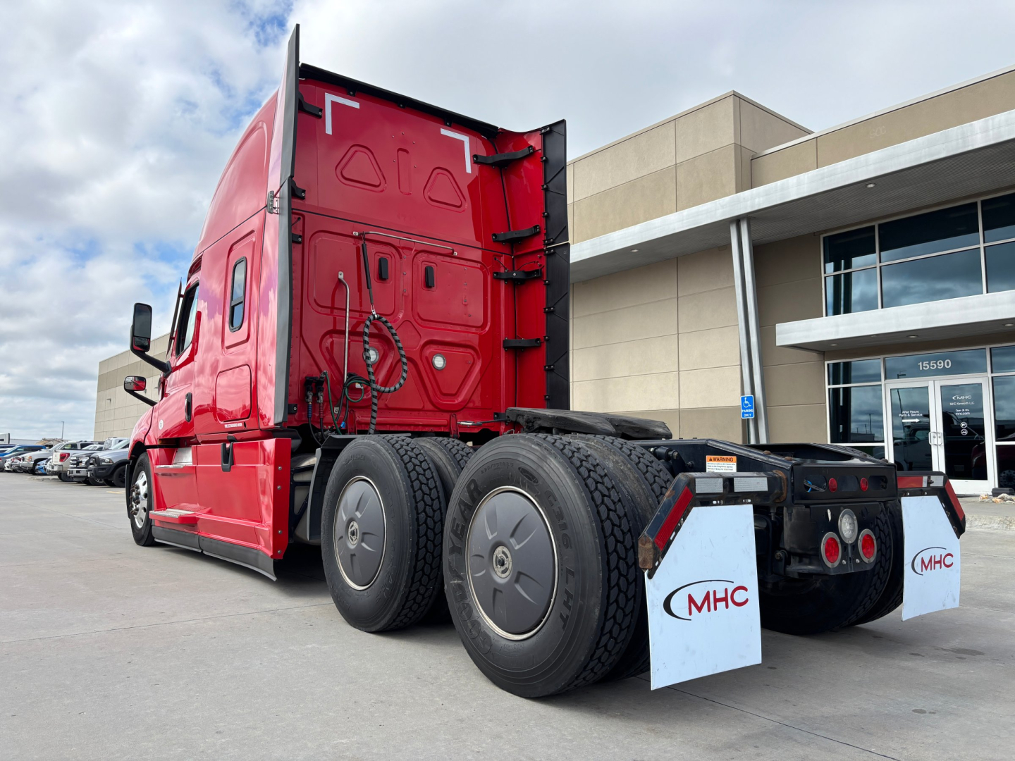 Truck rear view showing Freightliner PT126064ST sleeper tractor with dual rear axles and MHC mud flaps, red cab, dealership lot in background.