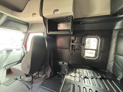 Truck cab interior showing storage compartments and sleeper area with window hatch and seat; cabin walls with a small closet-like box above and a metal floor plate.