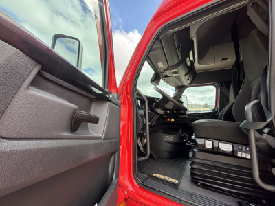 Truck cabin interior view from open driver door showing steering wheel, dashboard, pedals, and seat with red exterior.