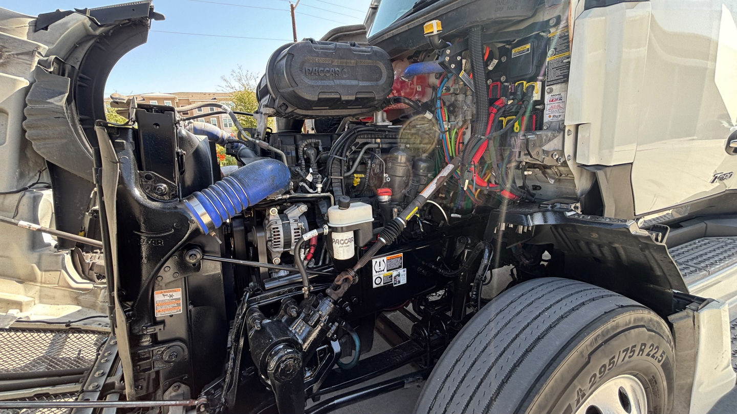 Kenworth T680 engine bay view showing various hoses, wiring, and components under hood; exposed intake, coolant reservoir, and plumbing.